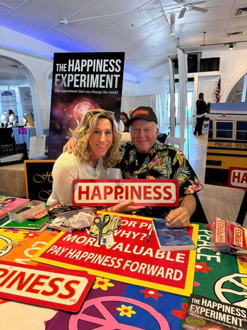 Gary King at an event table with a community member, surrounded by HAPPINESS signs and books