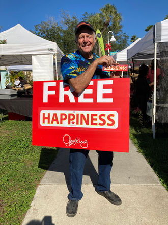 Gary King at an outdoor market holding a large red FREE HAPPINESS sign with his signature