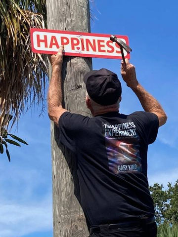 Gary King hammering a red HAPPINESS sign onto a wooden utility pole under a blue Florida sky