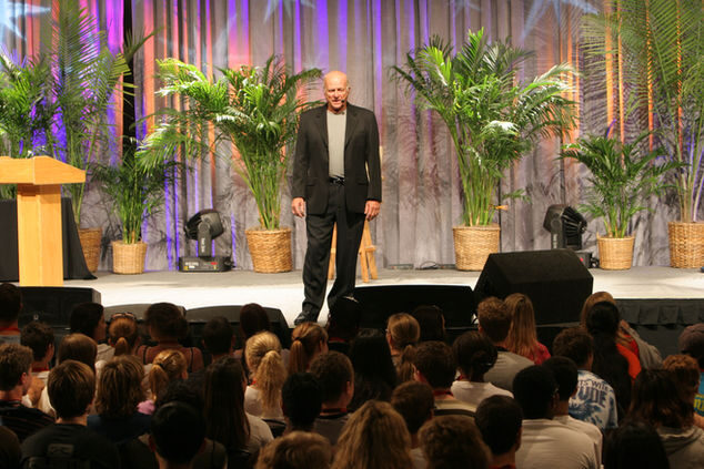 Gary King on a large stage addressing a packed auditorium of hundreds of young people, with palm plants and purple stage lighting