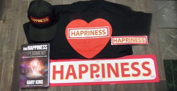 A flat-lay of Happiness Experiment merchandise: the book, a black t-shirt with a red heart and HAPPINESS text, a baseball cap, a small sign, and a full-size red metal HAPPINESS sign