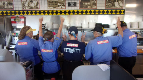 Five Waffle House workers at the counter with their backs turned, each wearing a blue uniform shirt with a HAPPINESS sign attached, arms raised in celebration